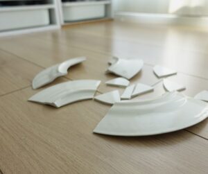Broken ceramic plate fragments on a kitchen floor in a quiet, orderly room. A symbolic image representing the invisible aftermath of domestic violence and trauma.
