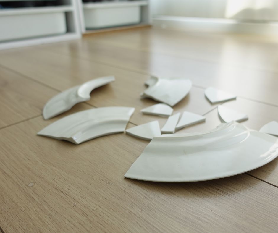 Broken ceramic plate fragments on a kitchen floor in a quiet, orderly room. A symbolic image representing the invisible aftermath of domestic violence and trauma.